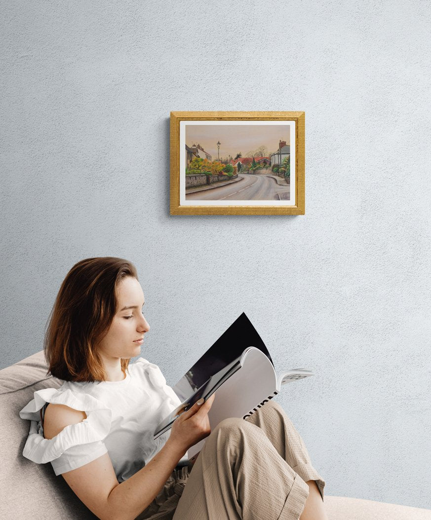 A woman sits on a light beige sofa reading a book, with a framed landscape artwork hanging on a textured light blue-gray wall above her.